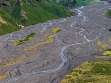 Örgülü bir nehir İzlanda 'da geniş kayalık bir vadi boyunca akar, koyu gri çökeltiyi örer. Vadiyi çevreleyen yemyeşil tepeler, canlı zıtlıklar yaratır..