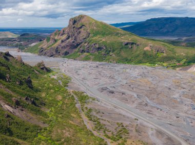 İzlanda 'da sarp kayalık yamaçlarla kaplı yeşil bir dağın havadan görünüşü çakıl düzlüklerinde örülmüş nehir kanalları ve parçalı bulutlu bir gökyüzünün altındaki uzak tepeler..