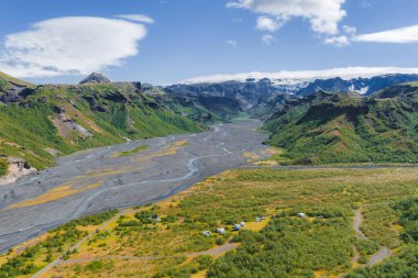 İzlanda 'da örülmüş bir nehir, engebeli dağlar, karlı zirveler, bir kamp alanı ve bulutlu parlak mavi bir gökyüzü olan yeşil bir vadinin havadan görünüşü..