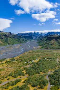 İzlanda 'da dolambaçlı bir nehir, engebeli dağlar, buzullar, dağınık çadırlar, araçlar ve parlak mavi gökyüzü olan yeşil bir vadinin havadan görünüşü..