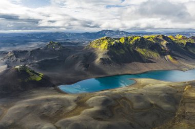 Karanlık volkanik araziler ve İzlanda 'da parçalı bulutlu bir gökyüzünün altındaki yosun kaplı tepelerle çevrili canlı mavi bir göl..
