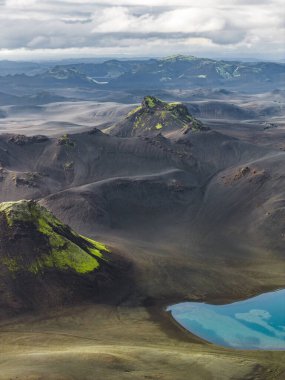 Yeşil yosun kaplı tepelerle İzlanda 'nın engebeli volkanik arazisinin havadan görünüşü ön planda turkuaz bir göl ve uzak sıradağlar..