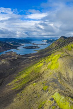 Yeşil yosun kaplı tepeler, volkanik arazi, adalı sakin bir göl ve dramatik sıradağlar içeren İzlanda dağlarının havadan görünüşü.
