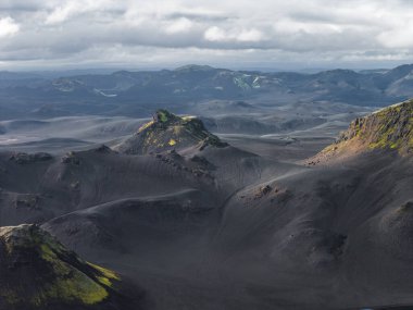 Parlak yeşil yosunları, engebeli tepeleri ve bulutlu gökyüzünün altında uzak dağları olan karanlık volkanik arazi dramatik bir İzlanda manzarası yaratıyor..