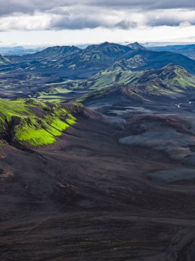 Karanlık toprak, canlı yeşil yosun kaplı tepeler ve dağınık ışık altında engebeli dağlarla İzlanda 'nın volkanik arazisinin dramatik hava manzarası..