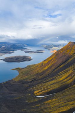 Renkli bir dağ yamacı, adalı sakin bir göl, karanlık volkanik arazi ve soluk bir gökkuşağı içeren İzlanda 'nın canlı bir hava görüntüsü..