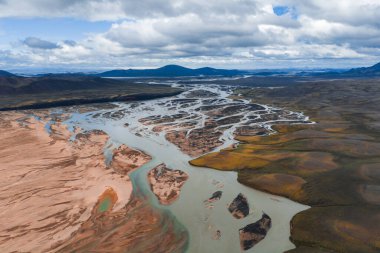 İzlanda 'da birbirine dolanmış nehir kanallarının hava görüntüsü, kırmızı kahverengi tortu, yeşil yosun, volkanik toprak ve uzak dağlarla çevrili..