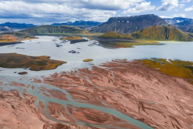 Bir göl, yosun kaplı tepeler ve bulutlu bir gökyüzü altında kar kaplı dağlar ile, kıpkırmızı kahverengi tortu boyunca akan örülmüş bir nehrin havadan görünüşü..
