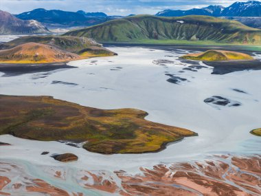 İzlanda 'nın çok renkli tepelerini gösteren hava manzarası, örülmüş buzullu nehirler, tortu desenleri ve bulutlu bir gökyüzünün altındaki kar tepeleri..