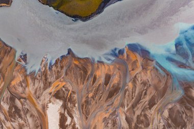 İzlanda 'da örülmüş bir nehir sisteminin hava görüntüsü, turuncu, kahverengi ve mavi renkte, manzaranın üstünde yeşil bitki örtüsü var..