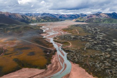 Volkanik kaya oluşumları, karlı dağlar ve parçalı bulutlu gökyüzü ile İzlanda 'nın renkli Landmannalaugar bölgesindeki dolambaçlı bir nehrin havadan görünüşü.
