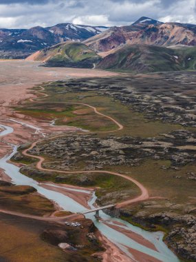 Buzul nehirlerinin dalgalı manzarası kayalık arazide dönüyor, küçük bir köprü, toprak bir yol, ve İzlanda 'da karlı zirveleri olan renkli tepeler..