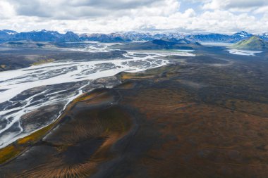 Karanlık toprak, turuncu ve yeşil renkler ve arka planda karla kaplı dağlarla İzlanda 'nın volkanik dağlarında kıvrılan örülmüş bir nehrin havadan görünüşü..