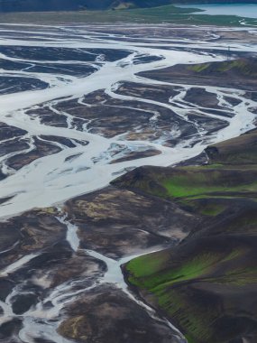 Örülmüş buzul nehirleri İzlanda 'da yeşil yosun kaplı tepelerle çevrili karanlık volkanik kumlar boyunca akar ve arka planda görünen bir göl vardır..
