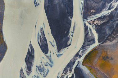 Örülmüş buzullar İzlanda 'daki karanlık volkanik kumların üzerinden akar, sağdaki turuncu ve yeşil bitki örtüsüne karşı sütlü beyaz su zıt düşer..