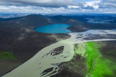 Canlı mavi bir göl, kıvrımlı bir nehir, yeşil yosun, yuvarlanan tepeler ve parçalı bulutlu bir gökyüzünün altındaki uzak dağların arasında yer alır..