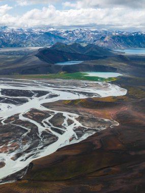 Yeşil bitki örtüsü, sakin bir göl ve İzlanda 'da karla kaplı dağlarla kaplı karanlık volkanik arazide kıvrılan örülmüş bir nehrin havadan görünüşü..