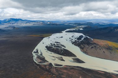 İzlanda 'daki karanlık volkanik arazide süzülen soluk bir buzul nehrinin hava manzarası, engebeli dağlar ve arka planda bulutlu gökyüzü..