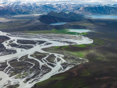 İzlanda 'da örülmüş bir nehir sisteminin havadan görünüşü, karanlık volkanik arazide yeşil yamalar, karla kaplı dağlar ve sakin bir göl..