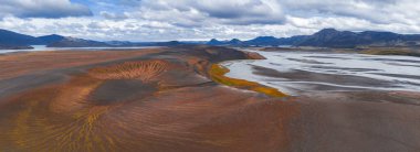 Kırmızımsı renkleri olan volkanik bir krater ve ovalarla çevrili sarı bitki örtüsü, dolambaçlı bir nehir ve parçalı bulutlu bir gökyüzünün altındaki uzak dağlar..
