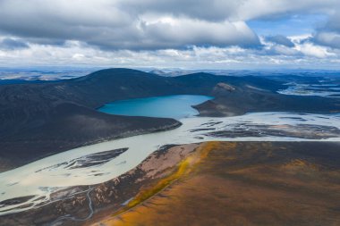 Karanlık volkanik tepelerle çevrili canlı mavi bir göl, engebeli arazide örülmüş bir nehir sistemi var. Topraksı tonlar ve sarı bitki örtüsü görünür..