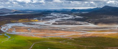 Örülmüş nehirler, yeşil otlar, volkanik topraklar ve parçalı bulutlu gökyüzünün altındaki uzak dağlarla çorak İzlanda topraklarında akıyor..
