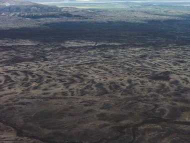 İzlanda 'da geniş bir volkanik arazi. Karanlık tepeleri, çöküntüleri ve uzak dağları var. Ufukta soluk bir su kütlesi ya da buzul görüntüsü beliriyor.