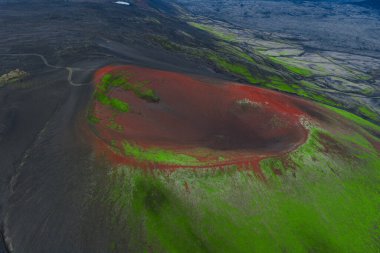 İzlanda 'daki volkanik bir kraterin hava görüntüsü. Siyah arazi, toprak yol ve uzak engebeli manzaralarla çevrili canlı kırmızı ve yeşil tonlarıyla..
