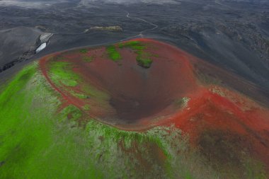 İzlanda 'da kırmızı ve yeşil yamaçlı bir volkanik kraterin havadan görünüşü, çorak arazi, uzak bir toprak yol ve arka planda kar..
