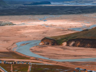Kayalık bir tepe, park halindeki araçlar, toprak raylar ve İzlanda 'daki uzak tepelerle dolambaçlı bir nehrin havadan görüntüsünü süpürüyor..