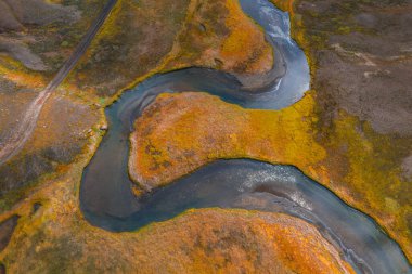 İzlanda 'da altın ve turuncu sonbahar bitkilerinin ortasında, solunda toprak bir yol ve arka planda sessiz bir arazi bulunan dolambaçlı bir nehrin hava görüntüsü..