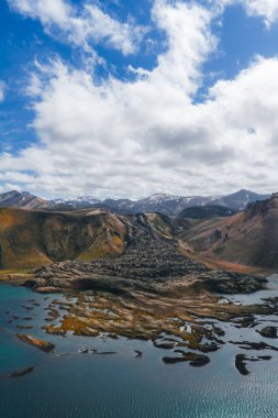 Çok renkli tepeler, kar tepeleri ve İzlanda 'da parçalı bulutlu bir gökyüzü ile çevrili, dingin mavi gölle buluşan volkanik bir lav tarlasının havadan görüntüsü..