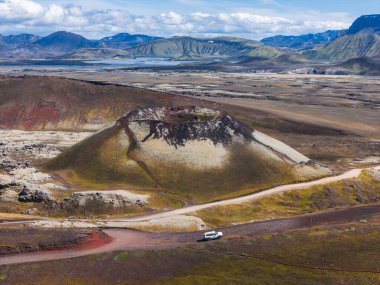 İzlanda 'da kırmızı, kahverengi ve yeşil bölgelerle çevrili bir volkanik krater. Beyaz bir araçla toprak bir yol kraterin etrafını sarmış tepeler ve yakınlarda bir göl var..