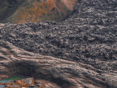 İzlanda 'daki engebeli volkanik arazi koyu lav tarlaları, toprak turuncu ve yeşil yamaçlar ve ön planda canlı yeşil ve turuncu bir havuz..