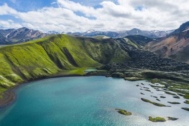 Turkuaz göl yosun kaplı tepeler ve volkanik arazilerle çevrili, arka planda kar kaplı dağlar parçalı bulutlu bir gökyüzünün altında..
