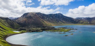 Yeşil yosunlarla çevrili canlı bir mavi göl ve İzlanda 'da parçalı bulutlu bir gökyüzünün altında kar kaplı tepeleri olan çok renkli dağlar..