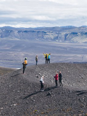 İzlanda 'da kara bir volkanik tepenin üzerinde bir grup duruyor. Etrafı çorak arazi, dramatik uçurumlar ve parçalı bulutlu bir gökyüzünün altındaki platolar ile çevrili..