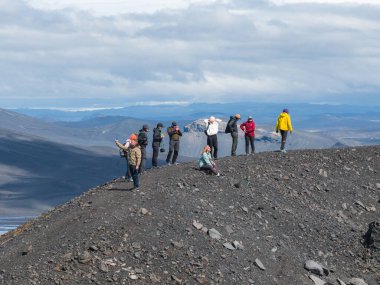 Açık hava giysileri içindeki insanlar, İzlanda 'da parçalı bulutlu bir gökyüzünün altında volkanik arazisi ve uzak buzulları olan kayalık bir tepede toplanıyorlar..