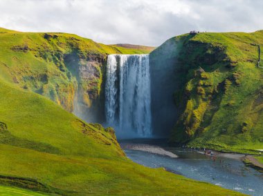 İzlanda 'daki Skogafoss şelalesi yeşil tepelerle çevrili yüksek bir uçurumdan akar. Nehrin aşağısındaki ziyaretçilerle şelaleden sis yükseliyor..