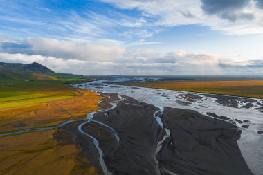 Örülmüş nehir kanalları İzlanda 'nın açık arazisindeki karanlık çökeltilerin arasından akar, yeşil bir yamaç ve bulutlu bir gökyüzünün altında altın günbatımı renkleriyle.