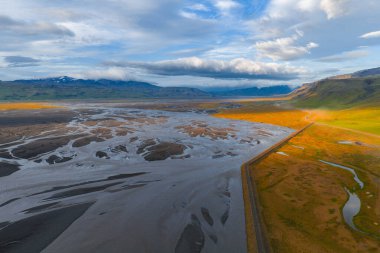 Karanlık çökeltilerle örülmüş nehir sistemi, düz bir yol, yeşil ve altın tarlalar, uzak karlı dağlar, ve İzlanda 'da günbatımı aydınlığı gökyüzü.