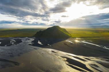 İzlanda 'nın düz bir manzarasından yeşil bir tepe yükseliyor, sarmal siyah kum nehir yatakları ve altın bir günbatımı gökyüzünün altında dereler..