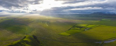 Gün batımında İzlanda 'nın yeşil ovalarının havadan görünüşü, güneş ışığı bulutların arasından akıyor, dolambaçlı bir yol, uzak dağlar ve dağınık binalar..