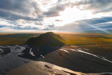Issız bir tepe İzlanda 'nın düz bir arazisinden yükselir, kıvrımlı siyah kum nehir yataklarıyla çevrilidir. Gün batımı ışığı yeşil ve topraksı tonları vurgular.