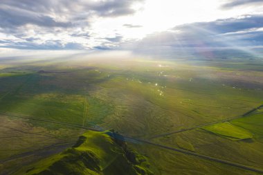 Gün batımında yeşil İzlanda manzarası, çimenli bir tepe, dağınık bulutlar, güneş ışınları, düz bir yol ve küçük su kütleleri..