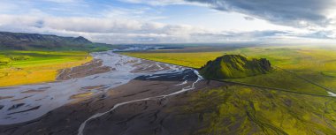 Düz bir araziden yükselen yeşil bir tepenin havadan görünüşü, örgülü bir nehir sistemi, canlı alanlar, volkanik toprak ve parçalı bulutlu bir günbatımı gökyüzünün altında bir yol..