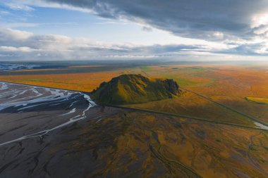 İzlanda 'daki düz turuncu ve yeşil tarlalardan yükselen yosun örtülü bir nehir ve günbatımı aydınlığı gökyüzünün altında düz bir yol.