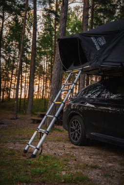 Siyah bir BMW X4 2025. Ormana park edilmiş, çatısı açık bir çadır ve merdiveni var. Etrafı uzun çam ağaçları ve güneş ışığıyla çevrili..
