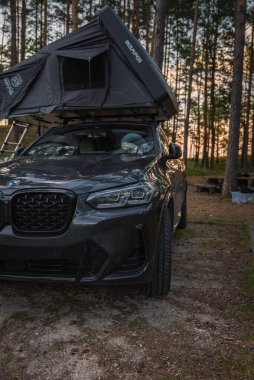 Bir BMW X4 2025 Kampa 'nın çatısı olan bir çadır ormanlık bir kamp alanında park edilmiş. Uzun çam ağaçları, bir piknik masası ve sıcak gün batımı ışığı görülebilir..
