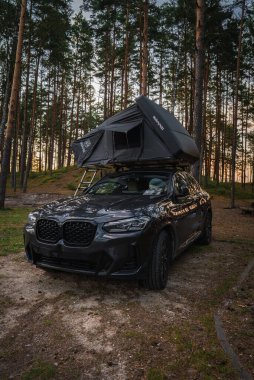 Siyah bir BMW X4 2025, çatısı açık bir çadır ve merdiveni uzun çam ağaçlarının arasına park etmiş, güneş ışığı ormanın içinden geçiyor..